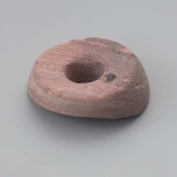 przęślik - Ujęcie z boku. Przęślik wykonano z łupku o różowej barwie. Zabytek jest uszkodzony, zachował się w około 50 %. Ma cylindryczny kształt z jednym zachowanym płaskim biegunem. W centralnej części znajduje się bardzo dokładnie wywiercony otwór przelotowy.