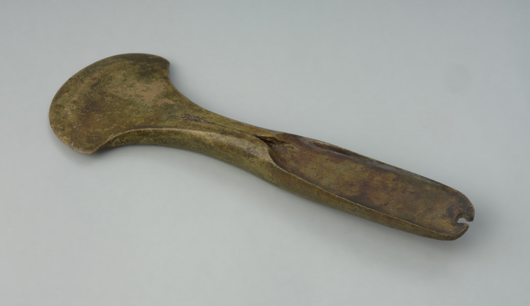 siekierka z piętką - Ujęcie z góry skosem do góry. przodu siekierki. Siekierka brązowa o zabawieniu złocistym z lejkowatą piętką i wąskim, łukowato zakończonym obuchem, na szczycie którego widnieje wyraźna wnęka. Ostrze zostało uformowane wachlarzowato.