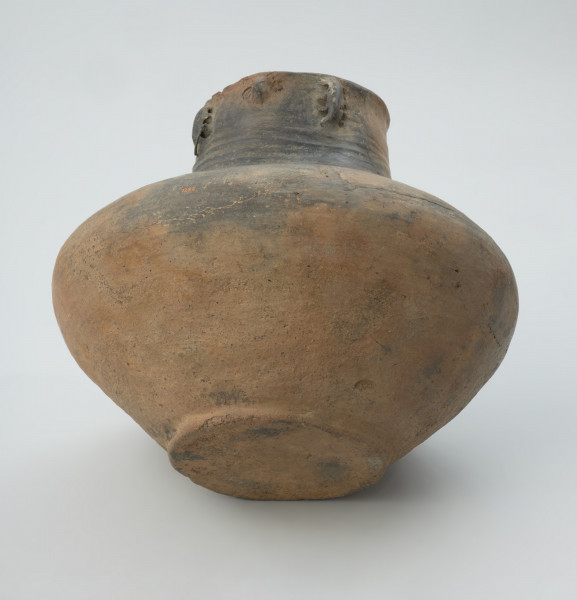 naczynie sepulklarne, urna twarzowa - Ujęcie skosem dołu i bocznej ścianki urny. Urna twarzowa z zaznaczonymi cechami twarzy ludzkiej, uszami ozdobionymi brązowymi kolczykami, ornamnetem rytym sugerującym naszyjnik oraz z pokrywką w kształcie kapelusza.