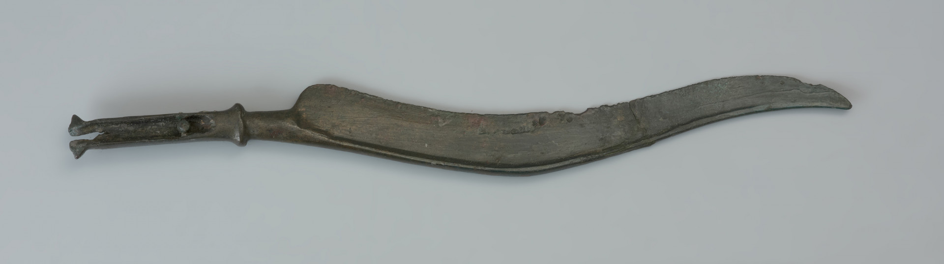 Nóż typu Pfatten - Ujęcie przodu noża z góry. Długi nóż wykonany z brązu ma smukłe esowato wygięte ostrze i wyraźnie zaznaczony tylec, przechodzący w sztabę do osadzania na niej rękojeści. Podstawa sztaby jest wyraźnie pogrubiona, jej szczyt natomisat uszkodzony. Profilowanie jej pozostałej części uzyskano poprzez charakterystyczne podniesienie brzegów. Przy podstawie zachował się niewielki nit służący do przytwierdzania rękojeści. Na zakończeniach po obu stronach sztaby widnieją dwa skrzydełka.