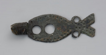 rozdzelnik naszyjnika z brązu - Ujęcie przodu z góry. Rozdzielnik naszyjnika z brązowej blaszki w kształcie ryby o długości 4,4 cm. W miejscu głowy znajduje się tulejka z widocznym łączeniem taśmy z przodu. Jej dalsza krawędź jest ścięta ukośnie po obu stronach. Tulejka zdobiona jest nacięciami skośnymi i prostopadłymi do jej przebiegu. Wzdłuż brzegów blaszki rozdzielnika znajduje się ornament z niewielkich dołków, natomiast wzdłuż jej dłuższej osi wykonano dwa otwory o nierównej średnicy. Dodatkowe dwa otwory znajdują się po obu stronach zwężenia przed 