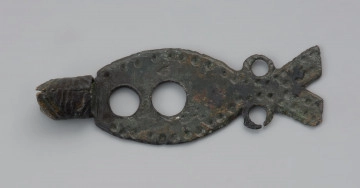 MNS/A/18327 rozdzelnik naszyjnika z brązu - Ujęcie przodu z góry. Rozdzielnik naszyjnika z brązowej blaszki w kształcie ryby o długości 4,4 cm. W miejscu głowy znajduje się tulejka z widocznym łączeniem taśmy z przodu. Jej dalsza krawędź jest ścięta ukośnie po obu stronach. Tulejka zdobiona jest nacięciami skośnymi i prostopadłymi do jej przebiegu. Wzdłuż brzegów blaszki rozdzielnika znajduje się ornament z niewielkich dołków, natomiast wzdłuż jej dłuższej osi wykonano dwa otwory o nierównej średnicy. Dodatkowe dwa otwory znajdują się po obu stronach zwężenia przed