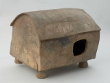 naczynie sepulklarne, urna domkowa - Ujęcie z przodu skosem do tyłu w prawą stronę. Urna gliniana w kształcie czworokątnego miniaturowego domu na czterech cylindrycznych filarach, z otworem drzwiowym zasłanianym ruchomą płytką i sekwencjami potrójnych linii rytych zdobiących dach.