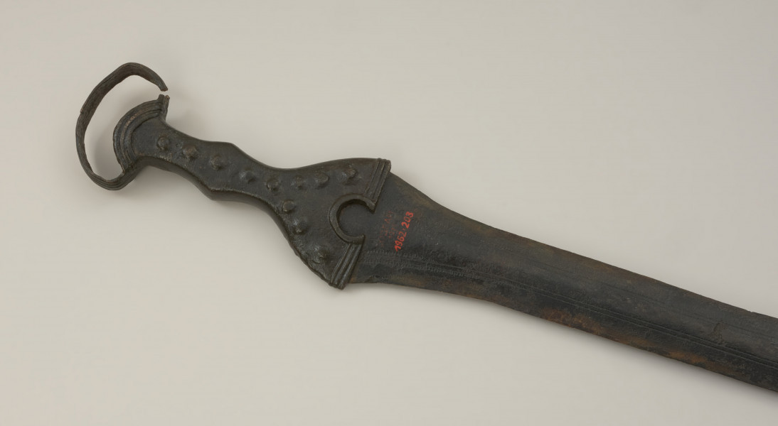 broń - Ujęcie rękojeści miecza od spodu w poziomie. Brązowy miecz posiada czworokątną rękojeść z nerkowatym pałąkiem oraz dzwonowaty jelec.