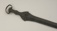broń - Ujęcie rękojeści miecza od spodu w poziomie. Brązowy miecz posiada czworokątną rękojeść z nerkowatym pałąkiem oraz dzwonowaty jelec.