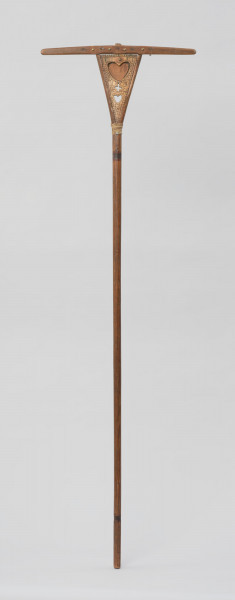 grabie - Ujęcie z przodu. Wykonane z drewna grabie pięciozębne. Długi drążek (rękojeść) rozszczepiona na końcu tworząc dwa ramiona. W miejscu, gdzie ramiona się rozchodzą wbity gwóźdź i owinięty wzmacniający konstrukcję sznurek.  Pomiędzy ramionami zamocowana, za pomocą gwoździ, wykonana z drewna trójkątna deseczka z dekoracją. Głównym elementem ornamentu są wycięte, ułożone pionowo nad sobą: dwa serca i  rozdzielający je krzyż. Powyżej górnego, większego serca wyryte litery: ET, poniżej data: 1874. Wzdłuż brzegów deseczki biegnie rząd trójkącików. Uzupełnieniem dekoracji jest kwiatek i dwa elementy geometryczne. Na rytach widoczne pozostałości farby w kolorze czerwonym i czarnym (ciemnozielonym?). Pośrodku górnego boku deseczki z dekoracją oraz na końcach ramion trzpienie, na które nasadzona jest poprzeczka  z zaokrąglonymi końcami. W poprzeczce wywiercone 5 otworów, w których mocowane są za pomocą ćwieków zęby.