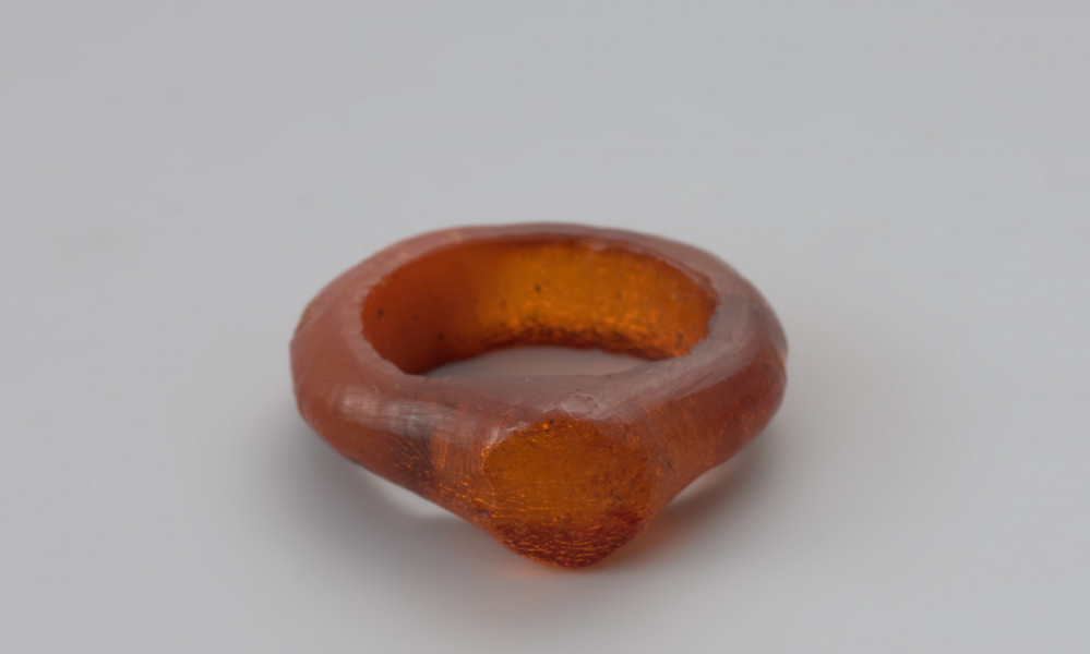 Jednoczęściowy pierścionek bursztynowy - Ujęcie z przodu w poziomie. Pierścionek bursztynowy z tarczką, jednoczęściowy, pomarańczowy, częściowo przejrzysty, częściowo mleczny. Jego otok jest w przekroju płaskokulisty i ma grubości ok. 0,4 cm. Powierzchnia zewnętrzna została wygładzona i nosi na sobie ślady obróbki, wewnętrzną natomiast pozostawiono nieobrobiona. Całość pokryta jest siatką drobnych pęknięć.