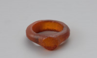 Jednoczęściowy pierścionek bursztynowy - Ujęcie z przodu w poziomie. Pierścionek bursztynowy z tarczką, jednoczęściowy, pomarańczowy, częściowo przejrzysty, częściowo mleczny. Jego otok jest w przekroju płaskokulisty i ma grubości ok. 0,4 cm. Powierzchnia zewnętrzna została wygładzona i nosi na sobie ślady obróbki, wewnętrzną natomiast pozostawiono nieobrobiona. Całość pokryta jest siatką drobnych pęknięć.