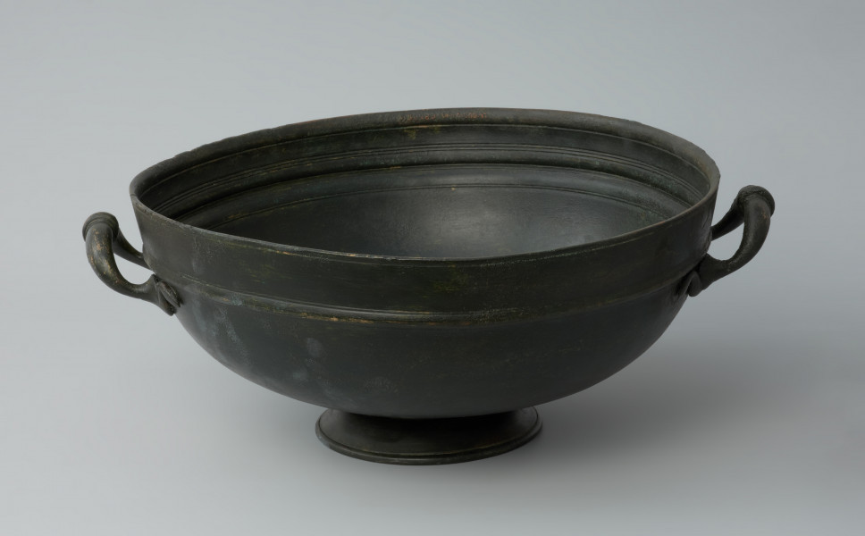 naczynie; Misa z brązu - Ujęcie z przodu. Misa z brązu na żebrowanej stopce. Na brzuścu znajduje się nieznacznie wypukły wałek.