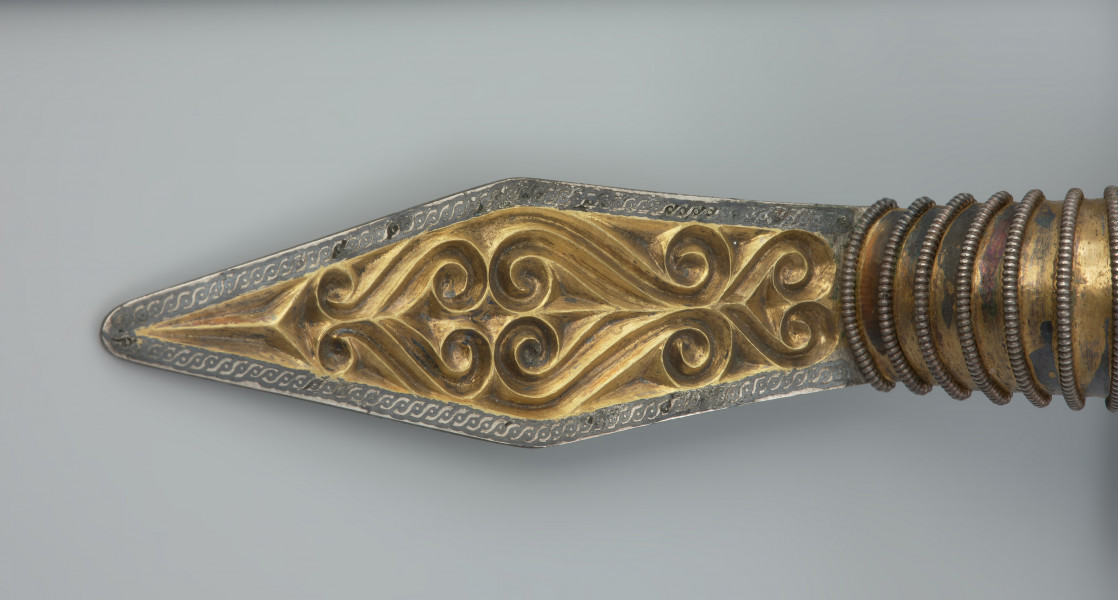 ozdoba; Srebrna, pozłacana fibula - Ujęcie detalu. Srebrna, pozłacana fibula z rombowato ukształtowaną długą nóżką, krótką pochewką i wysoko wysklepionym kabłąkiem. Zapinka ma główkę w postaci półkolistej tarczki nad sprężynką zaopatrzoną w 5 guzów wykonanych ze złotej taśmy.