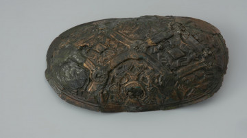 zapinka miseczkowata - Ujęcie z góry. Zapinka ma kształt wydłużonego owalu. Przedmiot jest częściowo uszkodzony bez zachowanej szpili. Powierzchnia fibuli jest bogato ornamentowana motywami zoomorficznymi. Na detal zdobniczy składa się osiem pól wyznaczonych przez krzyżujące się wstęgi. W punktach ich przecięcia znajdują się ślady po siedmiu wypukłych guzkach. Na sześciu z ośmiu pól znajdują się motywy zwierzęce bliskie skandynawskiej sztuce w stylu Borre. Na trzech z czterech zachowanych pól zlokalizowanych w narożnikach fibuli występują zwierzęta chwytające swoimi szponami ciała innych stworzeń. Dwa kolejne naprzeciwległe pola, usytuowane w środkowej części odpowiadają dwóm łukowato wygiętym w biodrach zwierzętom - być może są to konie z głowami skierowanymi w swoją stronę. Pozostałe dwa pola zawierają przedstawienia zwierzęcych masek z połączonymi ze sobą zaciśniętymi łapami.