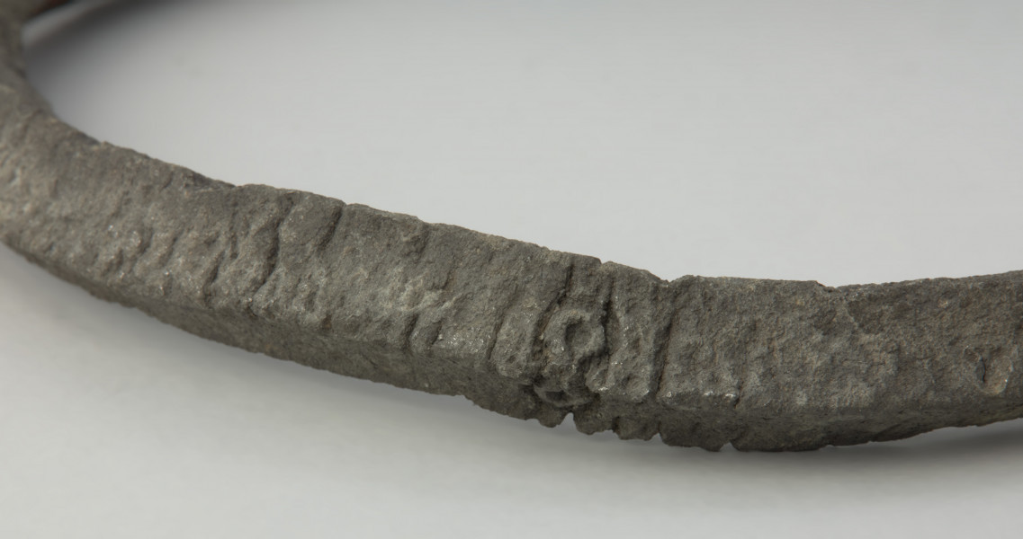 obręcz z czworograniastej sztaby - Ujęcie środkowego fragmentu obręczy. Srebrzystoszara obręcz, rozłamana na dwie części, wykonana została z czworograniastej cynowej sztaby. Przypomina kształtem naszyjnik obręczowy z ściętymi prosto i nieco pogrubionymi końcami oraz niewielkimi, nieregularnie rozmieszczonymi otworami. Zabytek jest zdeformowany, a jego powierzchnia popękana, co może świadczyć o wtórnym przepaleniu w ogniu.