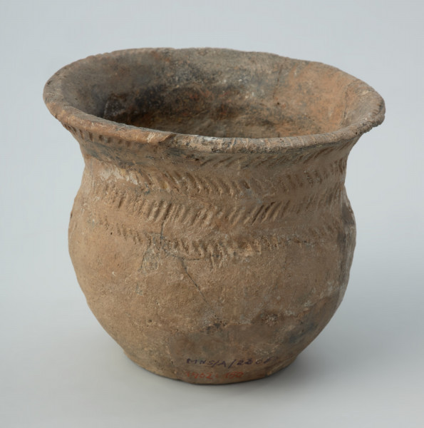 Naczynie gliniane - Ujęcie z tyłu; Przysadzisty puchar o szerokim wylewie, zdobiony na szyjce czterema dookolnymi rzędami ukośnych, podłużnych odcisków, które tworzą pasma złożone z dwóch szeregów odcisków układających się w motyw rybich ości. Ukośnymi nacięciami jest zdobiona także krawędź wylewu.