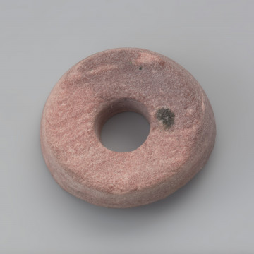 przęślik - Ujęcie z góry. Przęślik wykonano z łupku o różowej barwie. Zabytek jest uszkodzony, zachował się w około 50 %. Ma cylindryczny kształt z jednym zachowanym płaskim biegunem. W centralnej części znajduje się bardzo dokładnie wywiercony otwór przelotowy.