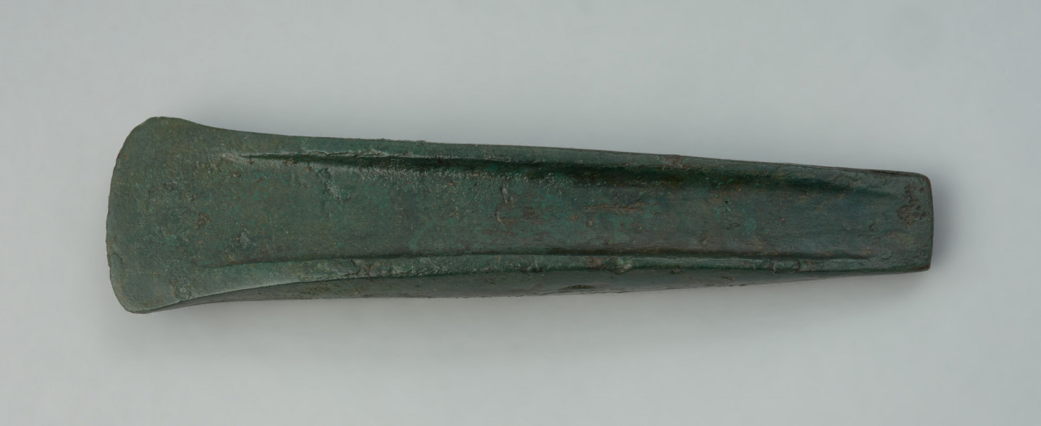 Smukła siekierka o trapezowanym kształcie - Ujęcie z góry w poziomie przodu siekierki. Smukła siekierka o trapezowanym kształcie, z dość wysoko podniesionymi brzegami biegnącymi nieprzerwanie od ostrza do końca prosto ściętego obucha. Ostrze jest nieznacznie poszerzone w stosunku do pozostałej części korpusu.