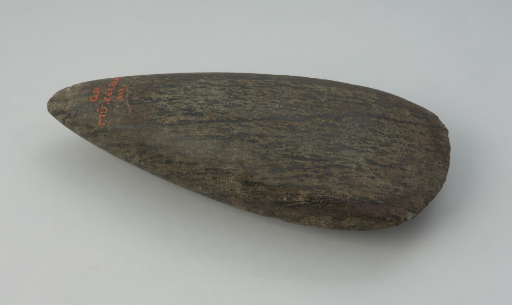 Siekiera kamienna - Ujęcie z dołu; Siekiera kamienna o owalnym przekroju poprzecznym, ze spiczastym obuchem oraz z nieco asymetrycznym, łukowatym ostrzem. Powierzchnia jest silnie zeszlifowana, równa i gładka. Wykonana jest z surowca skalnego o zielonkawej barwie z czarnymi pasmami.