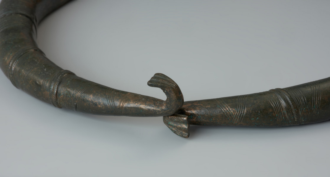 Dwuczęściowy naszyjnik obręczowy - Ujęcie detalu haczyków zapięcia. Zapinany haczykami naszyjnik z brązu, złożony z dwóch obręczy, zdobiony poprzecznym żebrowaniem oraz motywem zygzaka.