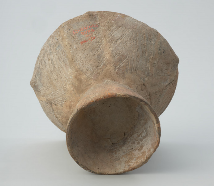 Naczynie gliniane - Ujęcie z dołu; Puchar na pustej nóżce, o klepsydrowatej formie, zdobiony na całej powierzchni ornamentem kreskowo-kłutym wykonanym dwuzębnym narzędziem: na czaszy – w układzie wiszących, wsuwanych kątów, które górą dochodzą do dookolnego pasma linii wykonanych tą samą techniką, a na nóżce – ukośnymi, zbieżnymi pasami linii. Przy krawędzi wylewu znajduje się zdobienie plastyczne w postaci czterech drobnych guzków