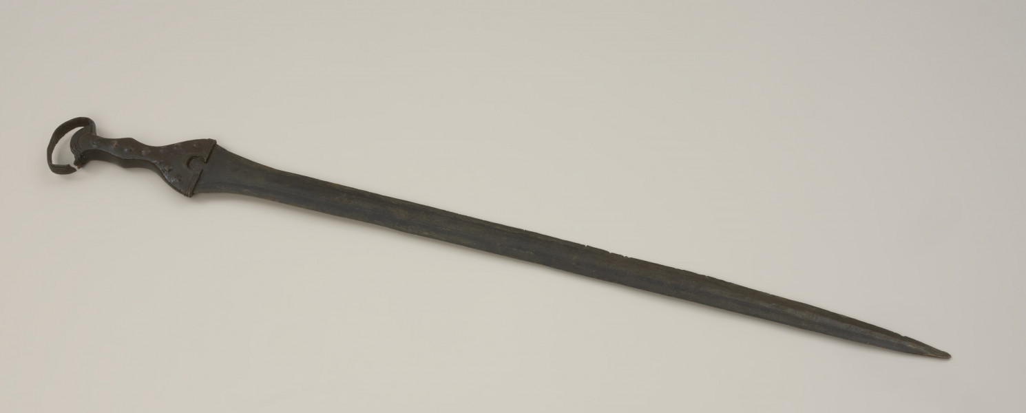 broń - Ujęcie przodu miecza. Brązowy miecz posiada czworokątną rękojeść z nerkowatym pałąkiem oraz dzwonowaty jelec.