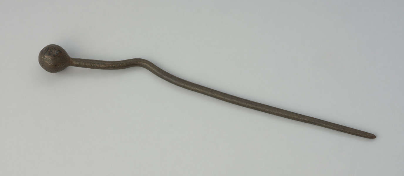 Szpila z brązu. - Ujęcie z góry. Szpila z brązu ze zdobioną główką motywem koncentrycznych okręgów i zygzaków oraz zakrzywionym trzonkiem, który również jest zdobiony pasmami dookolnych żłobień na odcinku między zakrzywieniem a główką.