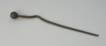 Szpila z brązu. - Ujęcie z góry. Szpila z brązu ze zdobioną główką motywem koncentrycznych okręgów i zygzaków oraz zakrzywionym trzonkiem, który również jest zdobiony pasmami dookolnych żłobień na odcinku między zakrzywieniem a główką.