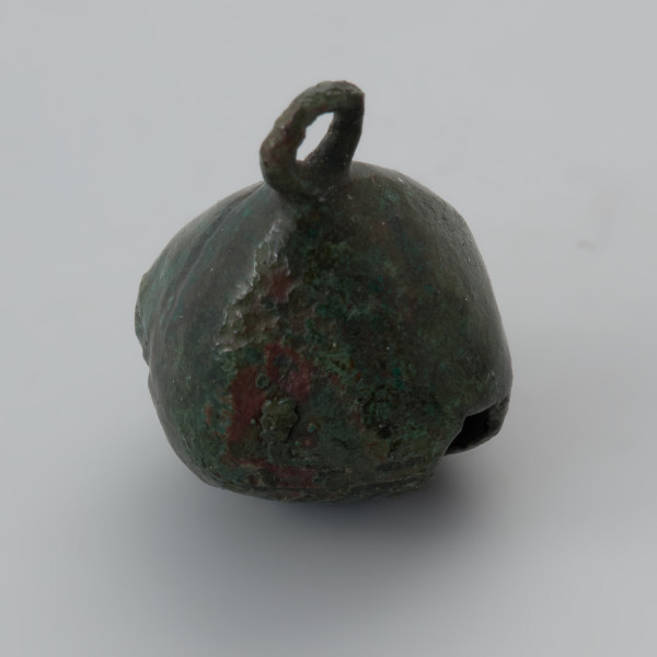 zawieszka dzwoneczkowata z Wolina - Ujęcie z góry przodu zawieszki skosem w lewą stronę. Zawieszka dzwoneczkowata wykonana z brązowej blachy, spatynowanej na ziolono. Korpus przypomina kształtem pustą w środku kroplę rozciętą u dołu na krzyż z jednym ramieniem dłuższym i szerszym od drugiego. Dodatkowo dolna powierzchnia jest zdobiona żłobkowaniem. W rzucie z góry zawieszka ma kształt zbliżony do rąbu, u góry posiada otwór z trójkątnego w przekroju drutu. W jej wnętrzu znajduje się brązowa kulka. Powierzchnia dzwoneczka jest lekko zniszczona, a w ściance znajduje się niewielki ubytek.