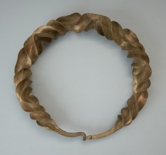 Naszyjnik wieńcowaty typu Wendelring - Ujęcie przodu z góry. Naszyjnik wieńcowaty charakteryzują szerokie, sfałdowane skrzydełka o ostrych krawędziach. Uzyskano je poprzez rozkuwanie i długotrwałe rozklepywanie pręta, pierwotnie o kwadratowym przekroju. Był skręcany wokół własnej osi przy kilkukrotnej zmianie kierunku skrętu. Czworokątne zakończenia naszyjnika ozdobione ornamentem punktowym wieńczy haczykowate zapięcie. Zabytek zachował się w dwóch częściach