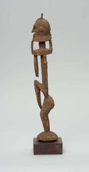 rzeźba - Ujęcie lewego boku. Drewniana, rzeźbiona postać znajdująca się w pozycji stojącej, przekrzywiona w lewą stronę. Głowa odcina się od szyi i tułowia prostą kreską. Ramiona proste. Ręce ułożone wzdłuż ciała. Nogi stosunkowo krótkie. Oczy, nos, usta wypukłe, broda w kształcie kryzy.