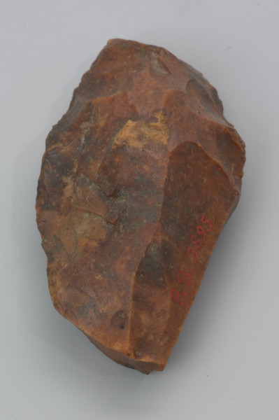 Mały ciosak rdzeniowy - ujęcie z przodu; Na zdjęciu ciosak rdzeniowy, narzędzie wykonano z krzemienia kredowego. Pokryte jest czerwonopomarańczową patyną, która jest efektem zalegania w środowisku bogatym w związki żelaza.