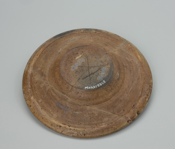 talerz - Ujęcie spodu. Wykonany na kole garncarskim z gliny o jasnej barwie ( z mała ilością tlenków żelaza). Dno słabo wyodrębnione przechodzi w niewysoki korpus o szerokim wlewie z wyraźnym kołnierzem. Szkliwo w tonacji żółtoszarej tylko na licowej powierzchni naczynia. Temat ornamentu wskazuje z dużym prawdopodobieństwem na to, że talerz był wyrobem jednostkowym wykonanym na zamówienie. Malatura przedstawia scenę rodzajowa: kobieta w niekompletnym stroju stoi pomiędzy dwoma mężczyznami. Jedna z tych postaci (większa) wyciąga ręce ku kobiecie , obok niej pies. Ponad postaciami , na kołnierzu, umieszczono napis: 