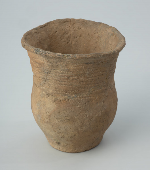 Naczynie gliniane - Ujęcie z przodu; Puchar o kształcie esowatym, zdobiony na szyjce dookolnymi pasmami odcisków sznura, złożonymi z 4 grup odcisków zwielokrotnionych linii, podkreślonym od dołu dookolną linią drobnych nakłuć. Powierzchnia nierówna, niestarannie wyrównywana, ze śladami po wypadających w trakcie wypału dodatkach w masie garncarskiej. Nad dnem wywiercony wtórnie otworek. Naczynie rekonstruowane i uzupełnione barwionym gipsem.