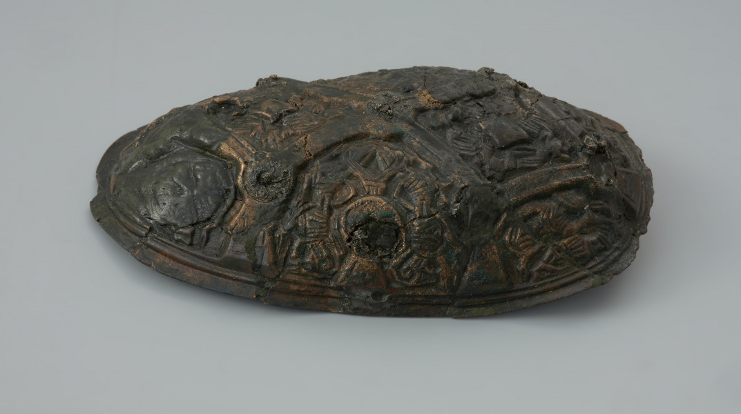 zapinka miseczkowata - Ujęcie prawego boku. Zapinka ma kształt wydłużonego owalu. Przedmiot jest częściowo uszkodzony bez zachowanej szpili. Powierzchnia fibuli jest bogato ornamentowana motywami zoomorficznymi. Na detal zdobniczy składa się osiem pól wyznaczonych przez krzyżujące się wstęgi. W punktach ich przecięcia znajdują się ślady po siedmiu wypukłych guzkach. Na sześciu z ośmiu pól znajdują się motywy zwierzęce bliskie skandynawskiej sztuce w stylu Borre. Na trzech z czterech zachowanych pól zlokalizowanych w narożnikach fibuli występują zwierzęta chwytające swoimi szponami ciała innych stworzeń. Dwa kolejne naprzeciwległe pola, usytuowane w środkowej części odpowiadają dwóm łukowato wygiętym w biodrach zwierzętom - być może są to konie z głowami skierowanymi w swoją stronę. Pozostałe dwa pola zawierają przedstawienia zwierzęcych masek z połączonymi ze sobą zaciśniętymi łapami.