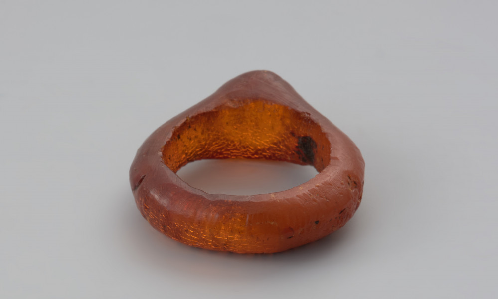Jednoczęściowy pierścionek bursztynowy - Ujęcie z tyłu w poziomie. Pierścionek bursztynowy z tarczką, jednoczęściowy, pomarańczowy, częściowo przejrzysty, częściowo mleczny. Jego otok jest w przekroju płaskokulisty i ma grubości ok. 0,4 cm. Powierzchnia zewnętrzna została wygładzona i nosi na sobie ślady obróbki, wewnętrzną natomiast pozostawiono nieobrobiona. Całość pokryta jest siatką drobnych pęknięć.