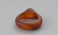 Jednoczęściowy pierścionek bursztynowy - Ujęcie z tyłu w poziomie. Pierścionek bursztynowy z tarczką, jednoczęściowy, pomarańczowy, częściowo przejrzysty, częściowo mleczny. Jego otok jest w przekroju płaskokulisty i ma grubości ok. 0,4 cm. Powierzchnia zewnętrzna została wygładzona i nosi na sobie ślady obróbki, wewnętrzną natomiast pozostawiono nieobrobiona. Całość pokryta jest siatką drobnych pęknięć.