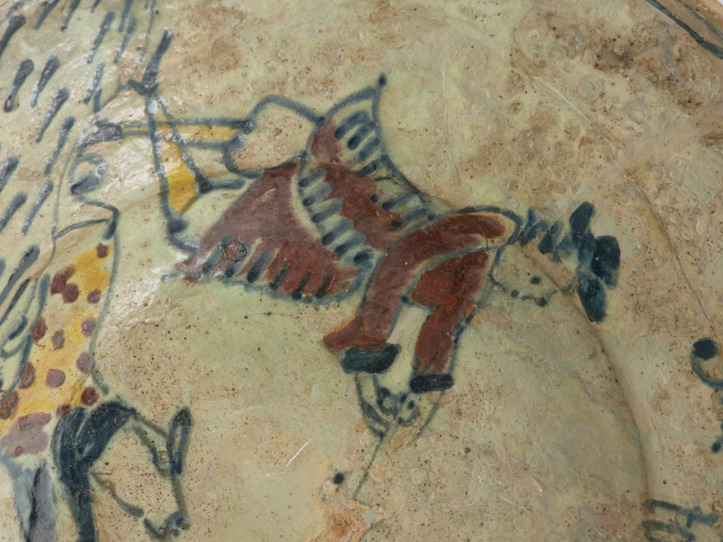 talerz - Ujęcie trzeciego fragmentu. Wykonany na kole garncarskim z gliny o jasnej barwie ( z mała ilością tlenków żelaza). Dno słabo wyodrębnione przechodzi w niewysoki korpus o szerokim wlewie z wyraźnym kołnierzem. Szkliwo w tonacji żółtoszarej tylko na licowej powierzchni naczynia. Temat ornamentu wskazuje z dużym prawdopodobieństwem na to, że talerz był wyrobem jednostkowym wykonanym na zamówienie. Malatura przedstawia scenę rodzajowa: kobieta w niekompletnym stroju stoi pomiędzy dwoma mężczyznami. Jedna z tych postaci (większa) wyciąga ręce ku kobiecie , obok niej pies. Ponad postaciami , na kołnierzu, umieszczono napis: 