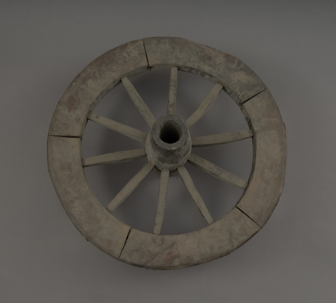koło - Ujęcie z góry. Replikę koła szprychowego wykonano na podstawie znalezisk z badań wykopaliskowych na Starym Mieście w Kamieniu Pomorskim. Jest produktem kołodzieja, inaczej stelmacha, rzemieślnika zajmującego się wyrobem wozów drewnianych. Poszczególne elementy koła zrobione są z różnych gatunków drewna.