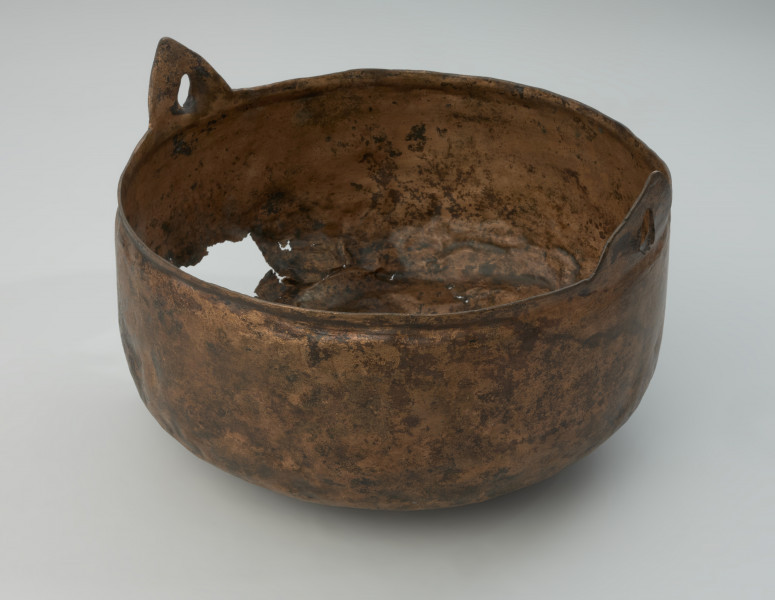 Kocioł ze stopu miedzi - Ujęcie z przodu skosem w lewą stronę. Kocioł ze stopu miedzi o owalnym kształcie z dwoma trójkątnymi uchami. Kocioł wykonano ze stopu miedzi. Jego kształt jest asymetryczny. W części górnej jest węższy i owalny, natomiast w części dolnej szerszy o zaokrąglonym dnie.