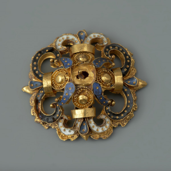 rozeta - Ujęcie z przodu. Ozdoba w kształcie ażurowej rozety. Wokół centralnie umieszczonego złotego guza rozmieszczone są cztery mniejsze guzy otoczone filigranem, płatki zdobione ciemnoniebieską emalią, a także woluty wypełnione czarną oraz białą emalią, spięte złotymi taśmami. Elementy emaliowane są inkrustowane regularnie rozmieszczonymi złotymi kropkami. Na odwrocie uszko służące do przyszywania rozet do strojów.