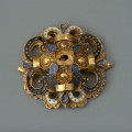 rozeta - Ujęcie z przodu. Ozdoba w kształcie ażurowej rozety. Wokół centralnie umieszczonego złotego guza rozmieszczone są cztery mniejsze guzy otoczone filigranem, płatki zdobione ciemnoniebieską emalią, a także woluty wypełnione czarną oraz białą emalią, spięte złotymi taśmami. Elementy emaliowane są inkrustowane regularnie rozmieszczonymi złotymi kropkami. Na odwrocie uszko służące do przyszywania rozet do strojów.