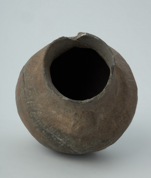 Naczynie gliniane - Ujęcie z dołu; Amfora o baniastym brzuścu i cylindrycznej szyjce z parą taśmowatych uch umieszczonych pomiędzy nasadą szyjki i dolną jej partią, zdobiona na szyjce i w górnej części brzuśca trzema dookolnymi pasmami pionowych i ukośnych słupków wykonanych w technice ściegu bruzdowego, rozdzielonych w połowie wysokości szyjki dookolną bruzdą oraz szeroką bruzdą ze śladami wygniatania palcami na przejściu szyjki w brzusiec. Naczynie zrekonstruowane i uzupełnione gipsem barwionym. Barwa od szarobrunatnej do brązowej.