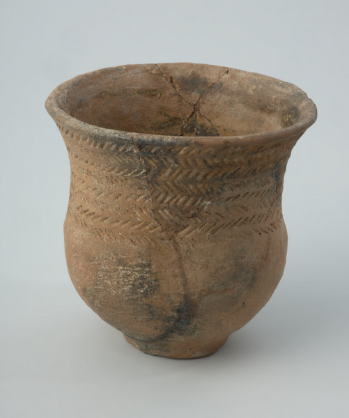 Naczynie gliniane - Ujęcie drugiego boku; Przysadzisty puchar o szerokim wylewie, zdobiony na szyjce czterema dookolnymi szeregami ukośnych, podłużnych odcisków, które tworzą pasma złożone z dwóch szeregów odcisków układających się w motyw rybich ości. Ukośnymi nacięciami jest zdobiona także krawędź wylewu.