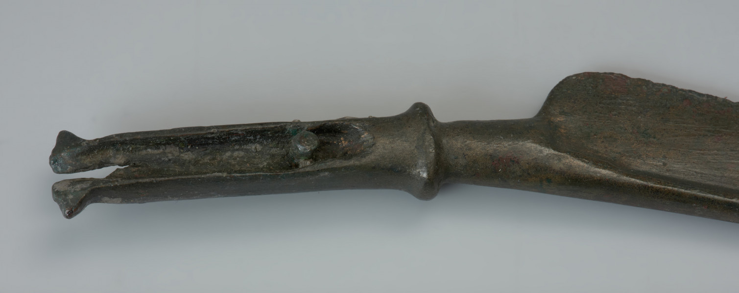 Nóż typu Pfatten - Ujęcie fragmentu rękojeści noża. Długi nóż wykonany z brązu ma smukłe esowato wygięte ostrze i wyraźnie zaznaczony tylec, przechodzący w sztabę do osadzania na niej rękojeści. Podstawa sztaby jest wyraźnie pogrubiona, jej szczyt natomisat uszkodzony. Profilowanie jej pozostałej części uzyskano poprzez charakterystyczne podniesienie brzegów. Przy podstawie zachował się niewielki nit służący do przytwierdzania rękojeści. Na zakończeniach po obu stronach sztaby widnieją dwa skrzydełka.