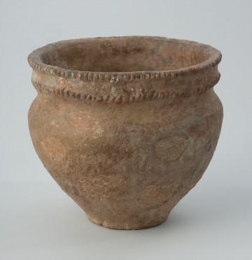 Naczynie gliniane - Ujęcie z przodu; Przysadziste naczynie pucharowate z lejkowatą szyjką i baniastym brzuścem, zdobione dwoma dookolnymi wałkami karbowanymi przy i poniżej brzegu. Powierzchnia chropowacona. Wyklejane na podstawie zachowanych fragmentów, zrekonstruowane dobarwionym gipsem