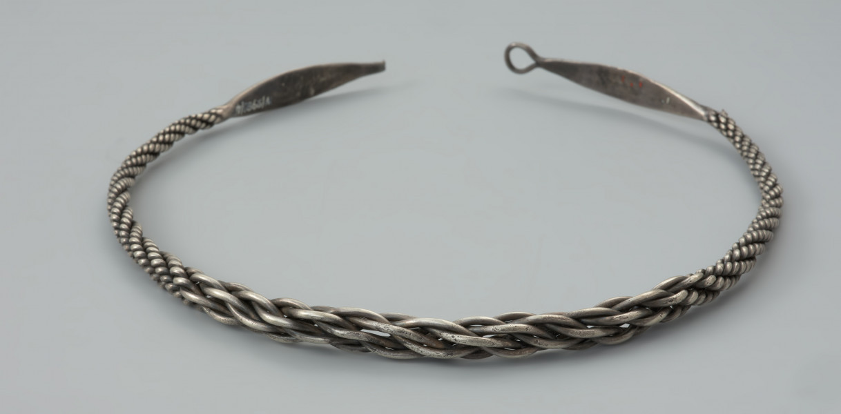 naszyjnik srebrny - Ujęcie przodu naszyjnika. Naszyjnik srebrny z czterech par skręconych drutów owijanych wokół siebie o grubości całkowitej od 0,4 do 1 cm. W części środkowej druty są skręcone zdecydowanie luźniej, co powoduje, że obręcz jest w tym miejscu grubsza. Oba końce obręczy są rozklepane w wydłużone owalne płytki zakończone uszkiem i haczykiem (fragment odłamany). Płytki są zdobione bordiurą z płaskich granulek wzdłuż obu krawędzi oraz dwoma rzędami trójkątów z trzema płaskimi granulkami w każdym z nich.