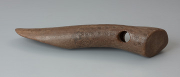 Topór zdobiony - Ujęcie topora z przodu w poziomie. Topór wykonano ze zrzutki poroża jelenia. W grubszej części przedmiotu znajduje się wywiercony otwór na stylisko. Powierzchnia topora w dużej mierze pokryta jest zdobieniem, tzw. motywem oczkowym, skłądającym się z kółeczka z zaznaczonym środkiem. Znajduje się tu także znak przypominający dużą literę 