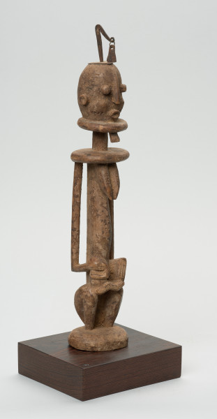 rzeźba - Ujęcie z przodu z prawej strony. Drewniana, rzeźbiona Postać z dzieckiem na kolanach. Siedzi w pozycji przykucniętej. Głowa i tułów nieproporcjonalnie duże w stosunku do nóg. Oczy, nos, usta, uszy mocno wypukłe. Dziecko ułożone na kolanach, zasłania twarz rękoma. Na głowie fragment żelaza.