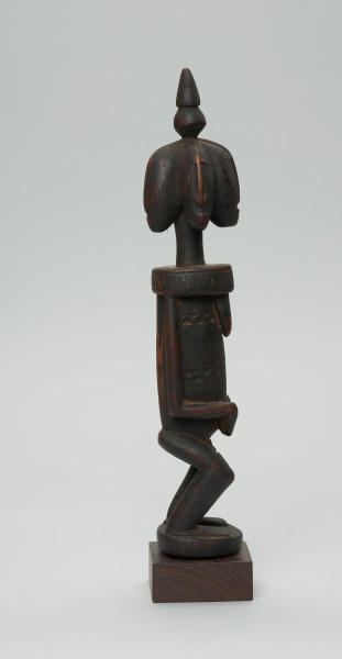 rzeźba - Ujęcie prawego boku. Drewniana, rzeźbiona postać z wyraźnie zaznaczonymi piersiami, a także z czterema głowami. Każda głowa zwrócona jest w inna stronę. Na czubku głowy stożkowata bryła. Twarze zaznaczone bardzo symbolicznie, nos w kształcie strzałki. Ramiona od szyi oddzielone prostą linią. Ręce przylegające do tułowia, dłonie złożone na łonie/podbrzuszu. Nogi krótkie, podkurczone w kolanach. Na brzuchu, na wysokości pępka dekoracyjne trójkątne nacięcia.