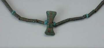 Kolia paciorków z wisiorkiem - Ujęcie fragmentu kolii w powiększeniu. Delikatna kolia złożona jest z niewielkich brązowych paciorków w kształcie rurek lub spiralek o średnicy 3 mm, separowanych drobnymi kolistymi paciorkami z zielonkawego szkła. Całość wieńczy wisior z brązu przypominający miniaturowy toporek o podwójnym ostrzu albo niewielkie śmigiełko o lekko wygiętych łopatkach. Wisior od strony wierzchniej jest wypukły, a od spodniej płaski. Środkowe zgrubienie zdobią poprzeczne żłobki.