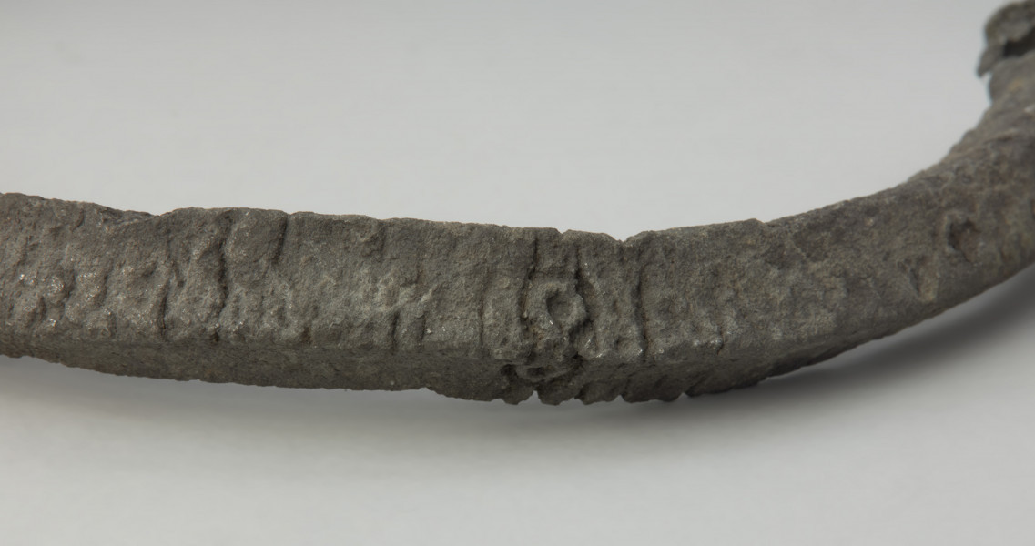 obręcz z czworograniastej sztaby - Ujęcie prawego fragmentu obręczy. Srebrzystoszara obręcz, rozłamana na dwie części, wykonana została z czworograniastej cynowej sztaby. Przypomina kształtem naszyjnik obręczowy z ściętymi prosto i nieco pogrubionymi końcami oraz niewielkimi, nieregularnie rozmieszczonymi otworami. Zabytek jest zdeformowany, a jego powierzchnia popękana, co może świadczyć o wtórnym przepaleniu w ogniu.