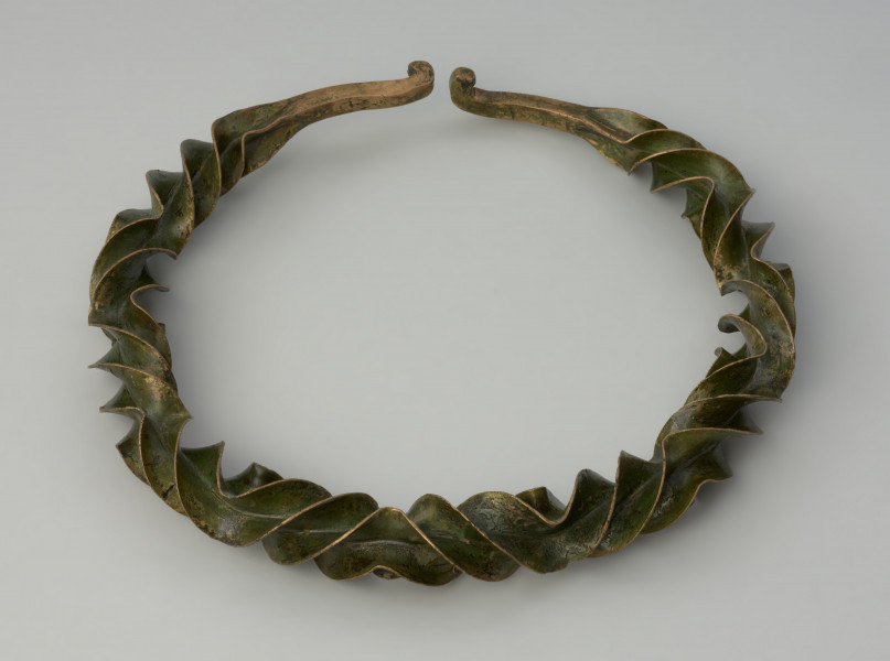 naszyjnik wieńcowaty typu Wendelring - Ujęcie z góry naszyjnika. Naszyjnik formą przypominający wieniec wykonano z brązowego, silnie rozklepanego na krawędziach, czworograniastego pręta o gwiaździstym przekroju, który skręcono (tordowano) wokół własnej osi zmieniając kilkukrotnie kierunek skrętu, uzyskując efekt wąskich, pofałdowanych płaszczyzn o ostrych krawędziach wzdłuż niemal całej długości obręczy. Końce ukształtowane zostały w haczyki służące zapięciu.